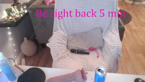 Media: A video of a person sitting in a white knit sweater, with a black vibrator and can of blue soda on a table. Text reads, \"Be right back 5 min.\