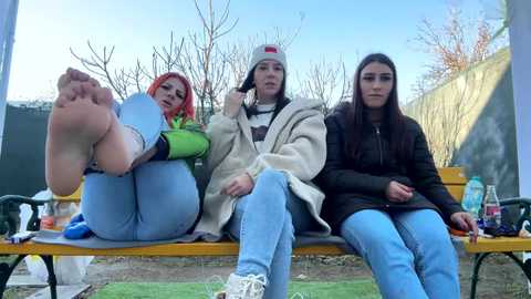 Media: Video of three young women with different skin tones and hairstyles sitting on a wooden bench in a park. They wear casual clothing, with one woman in a green hoodie, one in a beige blanket, and one in a black jacket.