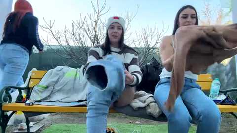 Media: Video of three young women in casual attire, sitting on a yellow bench in a park. One topless, one in a white beanie, one in a striped sweater.