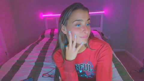 Media: Video of a young woman with light skin, long brown hair, wearing a red \"NUFU\" sweatshirt, smiling, in a dimly lit bedroom with a striped bedspread and pink LED lights.