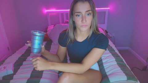 Media: Video of a young Caucasian woman with light skin, straight brown hair, and a slender build, sitting on a bed with a striped blanket. She wears a black t-shirt and white shorts. Neon pink lights illuminate the room.