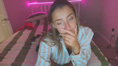 Media: Video of a young woman with light skin, long blonde hair, wearing a striped shirt, lying on a striped bed in a dimly lit room with pink LED strip lights.