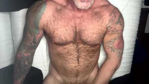 Media: Video of a muscular, hairy-chested man with tattoos on his arms and chest, lying on a white towel.