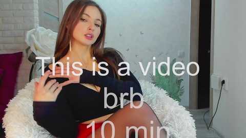 Media: Video of a young woman with long brown hair, wearing a black crop top and red skirt, sitting on a white shaggy rug, in a modern living room. Text overlay reads \"This is a video brb 10 min.\