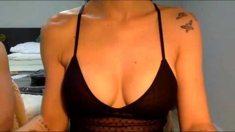 Media: Video of a light-skinned woman with a tattoo of a butterfly on her right shoulder, wearing a black halter top revealing ample cleavage. Background shows a bed with white sheets.