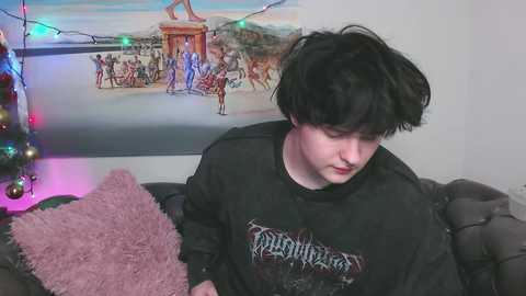 Media: Video of a young person with black hair, wearing a black Slayer t-shirt, sitting on a dark gray couch, surrounded by festive lights and a Christmas tree with ornaments.