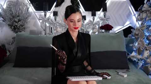 Media: Video of a woman with dark hair in a bun, wearing a black suit, red lipstick, and holding a cigarette, seated on a sofa with white decor and Christmas ornaments.