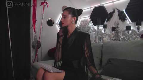 Media: Video of a slender, light-skinned woman with a medium build, Asian features, wearing black sheer gloves, and a black blazer, sitting on a gray couch in a dimly lit studio.