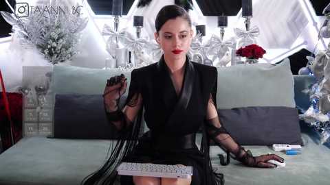 Media: Video of a poised woman with dark hair, red lipstick, and a black outfit, seated at a table with a laptop, surrounded by silver decor and a floral arrangement.