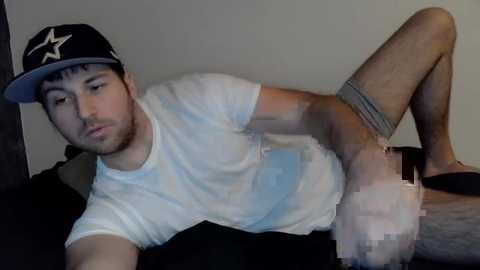 Media: Video of a young man with a beard, wearing a black cap and white t-shirt, masturbating on a bed.