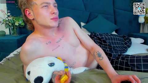 Media: Video of a shirtless young man with fair skin, tattoos, and short blonde hair, lying on a bed with a panda plushie, a plaid blanket, and teal pillows in the background.