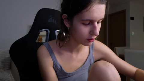 Media: Video of a young woman with fair skin, dark hair, and small breasts, wearing a gray tank top, sitting on a black gaming chair in a dimly lit room.