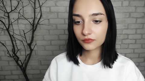 Media: Video of a young woman with straight, shoulder-length black hair, fair skin, and light makeup. She wears a plain white t-shirt. The background features a gray brick wall and a bare tree sculpture.