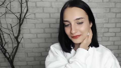 Media: Video of a young woman with straight black hair and light skin, wearing a white top, touching her cheek, against a white brick wall with a black decorative branch to the left.