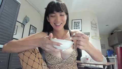 Video of a smiling Asian woman with long black hair, wearing a leopard-print top, holding a white bowl in a modern kitchen with white walls, framed pictures, and a fish tank.