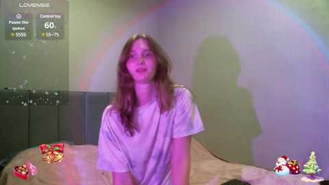 Media: Video of a young woman with long brown hair, wearing a white t-shirt, sitting on a bed in a dimly lit room with a rainbow projected on the wall.