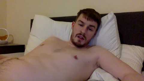 Media: A video of a nude, fair-skinned, muscular man with short brown hair and a trimmed beard, lying on a bed with white pillows and a dark headboard. The background is a simple beige wall.