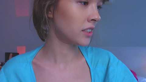 Media: Video of a young woman with short brown hair, fair skin, and a small nose piercing, wearing a light blue shirt, gazing off to the side. Background features a soft blue wall with subtle red lighting.