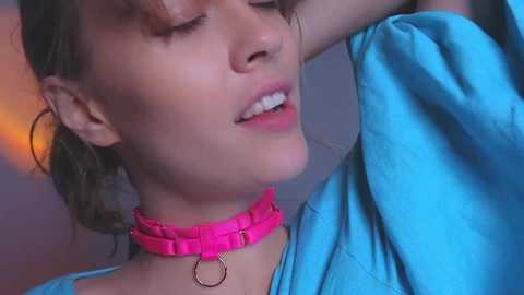 Media: Video of a young woman with light skin and dark hair, wearing a pink choker necklace and a blue shirt, posing with her eyes closed, possibly in a bathroom.