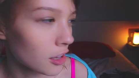 Media: A video of a young woman with light skin, dark hair, and blue eyes, wearing a pink tank top, biting her lower lip while indoors with a dimly lit room featuring a warm, orange lamp.