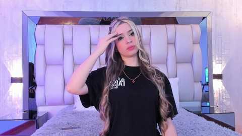 Media: Video of a young woman with long, wavy blonde hair, wearing a black T-shirt with white text, standing in a modern, minimalist bedroom with a tufted white headboard and soft lighting.