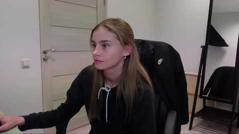 Media: Video of a young Caucasian girl with straight, light brown hair, wearing a black hoodie, sitting in a chair. She looks thoughtful. Background includes a white door, a black backpack, and a black chair.