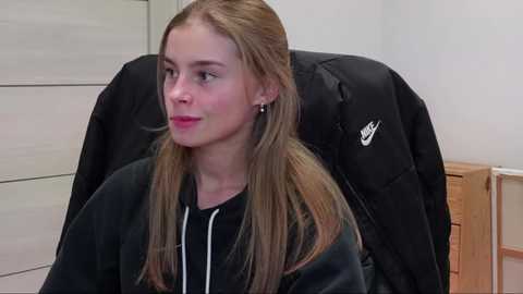 Media: Video of a young Caucasian woman with light brown hair and fair skin, wearing a black hoodie, sitting in a chair with a black Nike jacket draped over it in a minimalistic room with light-colored walls.