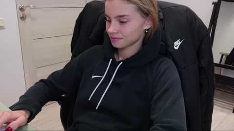 Media: Video of a young Caucasian woman with light skin, brown hair, and a black Nike hoodie, sitting in a black office chair, looking at a computer screen in a modern office setting.