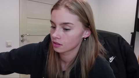 Media: Video of a young, fair-skinned woman with long, straight blonde hair, wearing a black Nike sweatshirt, seated in a chair, looking down, with a door and light switch visible in the background.