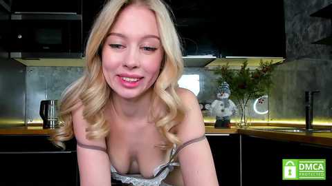 Media: A video of a smiling, blonde woman with medium breasts, wearing a white lace bra, in a modern kitchen with black cabinets, stainless steel appliances, and a metallic backsplash.