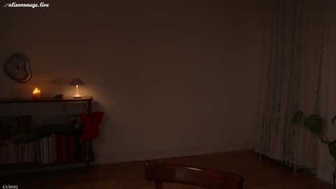 Media: A dimly-lit, minimalist living room photo with beige walls, a wooden table, and a bookshelf with books and a red towel. A wall clock and a lamp with a red shade provide soft light.