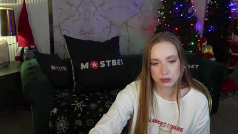 Media: Video of a young woman with long blonde hair, wearing a white sweater, sitting on a dark green couch adorned with \"MST3K\" pillows in a festive, dimly lit living room with a decorated Christmas tree and holiday decorations.