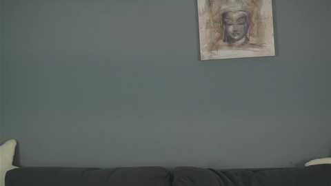 Video of a muted green wall featuring a framed, abstract painting of a serene, bearded face in earthy tones. Below, a black sofa with white cushions.