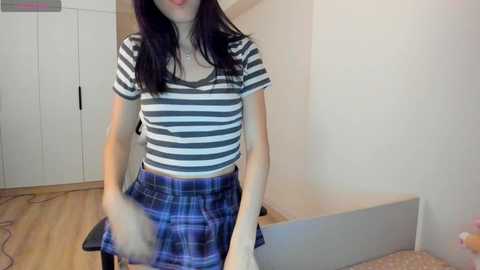 Media: Video of a young woman with long black hair, wearing a striped shirt and plaid skirt, sitting on a wooden floor in a simple room with white walls and minimal furniture.