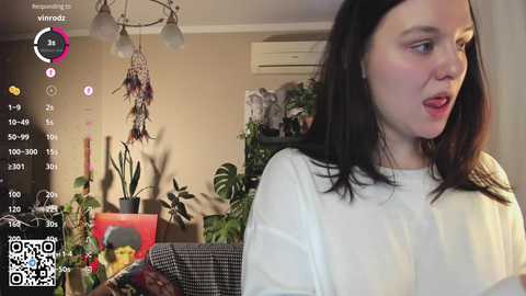 Media: Video of a woman with straight, dark hair, wearing a white sweater, in a cozy living room with green plants, hanging dreamcatchers, and a QR code overlay.