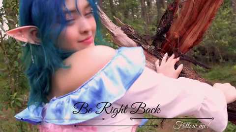 Media: Video of a young woman with teal hair and elf ears, wearing a tie-dye off-shoulder top and white pants, lying on a fallen tree in a forest. Text reads \"Be Right Back\" and \"Follow me!\