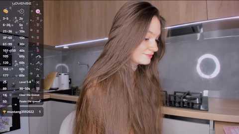 Media: Video of a young Caucasian woman with long, straight brown hair in a modern kitchen. She has fair skin and a slim build. The background features light wooden cabinets, a stainless steel stove, and a glass backsplash.