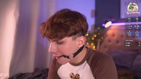 Media: Video of a short-haired, fair-skinned woman in a brown and white T-shirt, wearing a black ball gag and sitting on a bed. Background shows a dimly lit room with a TV screen displaying streaming stats.