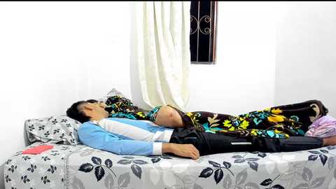 Media: Video of an Asian man lying on a bed with a floral-patterned duvet, wearing a blue and white shirt, black pants, and black shoes, in a simple, white-walled room with a barred window.