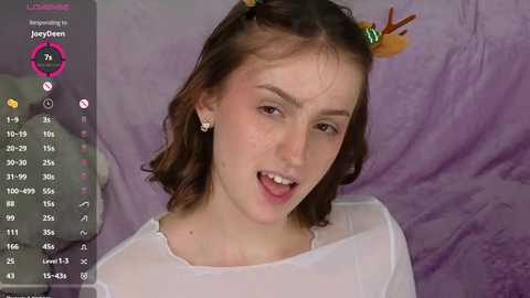 Media: Video of a young woman with light skin, brown hair, and a playful expression. She wears a sheer white top and colorful hair accessories against a lavender backdrop.