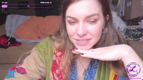 Media: Video of a woman with long, brown hair and fair skin, wearing a colorful, patterned cardigan, smiling and making a peace sign. She sits on a beige couch with a messy background.