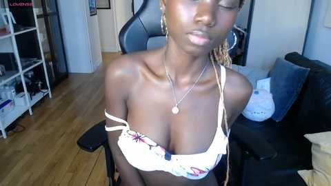 Media: Video of a Black woman with dark skin, bra straps off, revealing ample cleavage in a white lace bra, sitting on a black chair in a modern room.