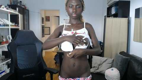 Media: Video of a slender, dark-skinned woman with braids, wearing a white crop top and floral panties, sitting in a cluttered living room with gaming setup.