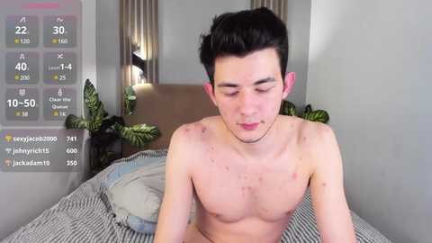 Media: A video of a young, shirtless man with fair skin, black hair, and a thin physique, sitting on a bed with striped sheets, in a modern bedroom.