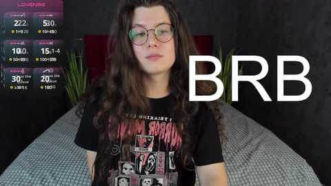 Media: Video of a young woman with long brown hair, wearing glasses and a black Nirvana t-shirt, sitting on a bed with a dark gray patterned blanket. A digital clock and \"BBR\" logo are overlaid.