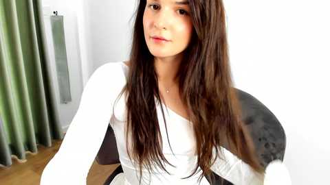 Media: Video of a young woman with long brown hair, wearing a white long-sleeve top, seated in a modern gray chair. Background features white walls and green curtains.