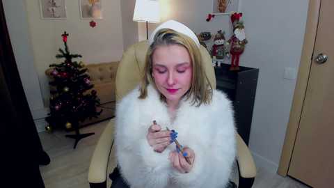 Media: Video of a blonde woman in a white fur coat and white hat, seated in a beige armchair, holding a blue toy. Christmas decorations adorn the background.