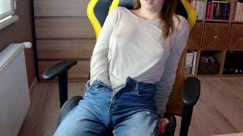 Media: Video of a young Caucasian woman with light skin and brown hair, sitting in a yellow and black gaming chair, wearing a sheer white top and high-waisted jeans. The room has wooden paneling, a radiator, and bookshelves in the background.