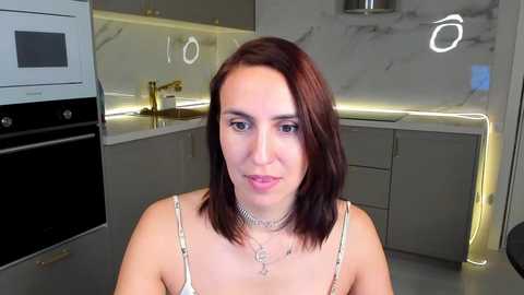 Media: Video of a woman with shoulder-length auburn hair, wearing a white spaghetti-strap top and a silver necklace, in a modern kitchen with white countertops, grey cabinets, and stainless steel appliances.