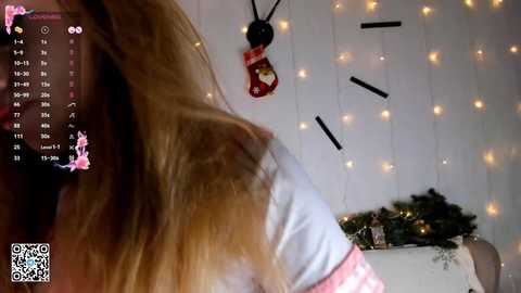 Media: Video of a blonde woman with long hair, wearing a white shirt, in a cozy room with string lights, a Christmas decoration, and a calendar.